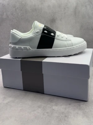 Valentino vita sneakers med svart detalj - Snygga vita sneakers från Valentino med en bred svart läderdetalj över sidan och coola nitar på hälen. Skorna har en chunky vit sula med mönstrad undersida och klassisk snörning. Perfekta för dig som vill ha en clean men ändå unik look. 