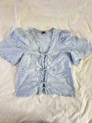 Ljusblå spetsblus från Vero Moda - Söt ljusblå blus från Vero Moda med broderade detaljer och spets. Blusen har korta puffärmar och knytning framtill med tre rosetter. Materialet är lätt och luftigt, perfekt för sommarens alla tillfällen.