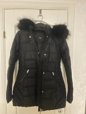 Svart dunjacka med fuskpäls - Snygg svart dunjacka från Rockandblue med figurnära passform och stor huva med svart päls. Jackan har dragkedjor på fickorna, quiltade detaljer på sidorna och en bröstficka med dragkedja. I princip helt oanvända nypris ligger på ca 4500.