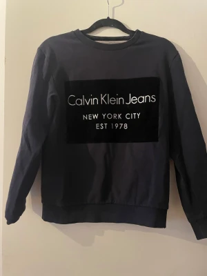 Mörkblå sweatshirt Calvin Klein Jeans - Mörkblå sweatshirt från Calvin Klein Jeans med stor mocca logga och texten 'New York City EST 1978' på bröstet. Tröjan har rund halsringning och ribbade muddar vid ärmslut och nederkant. Perfekt för en avslappnad och stilren look.