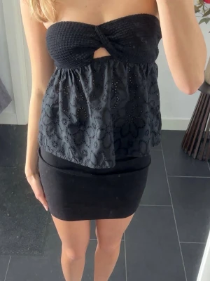 Svart bandeau tubtopp med broderi - Snygg svart bandeau tubtopp med broderade blommor och liten cut-out framtill. Toppen är axelbandslös och har en luftig passform nedtill. Perfekt för dig som vill ha en trendig och unik topp till sommaren.