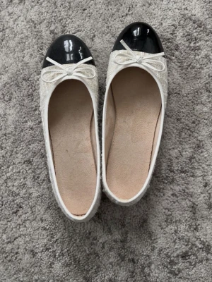 Vita ballerinaskor med svarta tår - Snygga vita ballerinaskor med glansig svart tå och dekorativ rosett framtill. Skorna har rundad form och ovandel i tyg med en lätt struktur. Perfekta för en stilren och klassisk look.