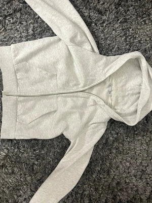 Ljusgrå zip hoodie från H&M - En stilren ljusgrå hoodie från H&M med dragkedja framtill och två stora fickor. Hoodien har en klassisk huva och ribbade muddar vid ärmslut och nederkant. Tillverkad i mjukt bomullsmaterial som är skönt att ha på sig.