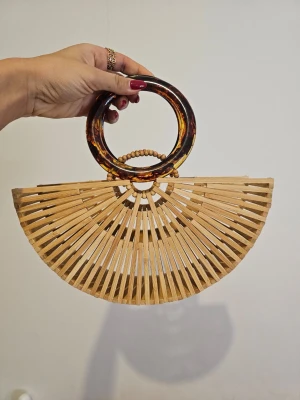 Halvmåneformad bambuväska med ringhandtag - Säljer en unik handväska i halvmåneform gjord av bambu med coola runda handtag i sköldpaddsmönstrad plast. Väskan har ett öppet, luftigt design och är perfekt för dig som gillar boho-stil och naturliga material.