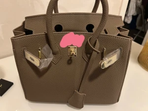 Beige handväska med gulddetaljer - Snygg beige handväska i strukturerat skinn med klassisk fyrkantig form och två rejäla handtag. Väskan har guldfärgade metalldetaljer och ett elegant lås framtill. Perfekt storlek för att få plats med allt du behöver och ger en lyxig vibe! Har logo storlek 20. STICKERS E FORTFARANDE PÅ