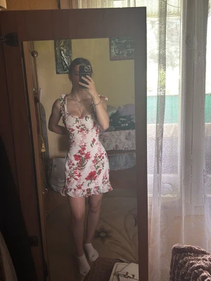 Vit blommig kortklänning - Super söt vit kortklänning med röda blommor, volangkant nertill och knytband på axlarna. Klänningen är ärmlös, perfekt för sommarens dagar. Materialet känns lätt och luftigt. Säljs då den tyvärr är för stor för mig.