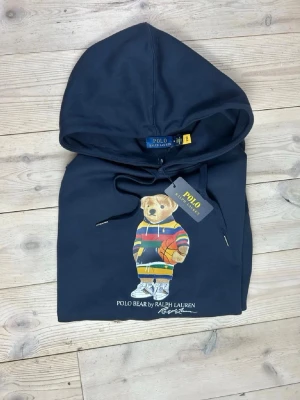 Mörkblå hoodie Polo Bear Ralph Lauren - Snygg mörkblå hoodie från Ralph Lauren med Polo Bear-motiv på bröstet. Björnen har färgglad tröja och sneakers, vilket ger ett coolt och lekfullt intryck. Hoodien har dragsko i huvan och är tillverkad i mjukt bomullsmaterial.