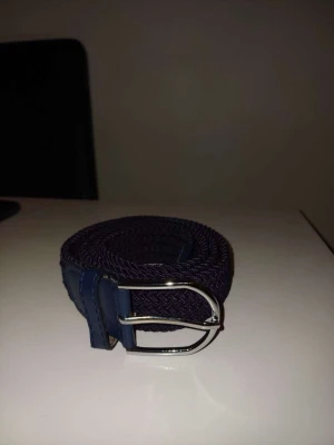 Mörkblått flätat bälte med silverdetalj - Snyggt mörkblått flätat bälte med silverfärgat spänne och blåa skinn-detaljer vid ändarna. Perfekt accessoars för att lyfta din outfit och passar till både jeans och chinos. Enkel att justera tack vare det flexibla materialet.