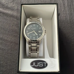 JUST silverfärgad klocka med länkarmband - Stilren klocka från JUST med silverfärgat länkarmband i metall och svart urtavla. Klockan har tydliga siffror, datumvisning och klassisk design som passar till många olika stilar. Perfekt accessoar för dig som gillar enkel och tidlös look (oanvänd) 