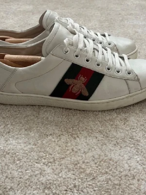 Gucci Ace vita sneakers med bi - Gucci Ace sneakers i vitt läder med klassiska gröna och röda ränder på sidan samt broderad guldfärgad bi. I bra skick och priset kan sänkas.