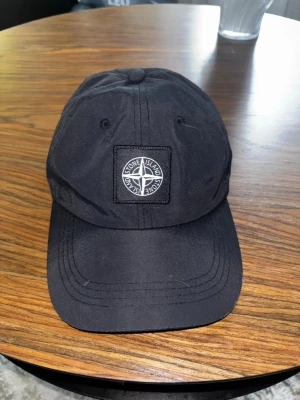 Svart keps från Stone Island - Snygg svart keps från Stone Island med klassisk böjd skärm och justerbart band bak. Framtill sitter en broderad patch med Stone Island-loggan i vitt och svart. Kepsen är tillverkad i ett slitstarkt tyg som ger en clean och stilren look.