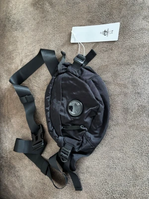 Svart C.P company crossbody-väska - Svart crossbody-väska från C.P company med justerbara remmar och spänne. Väskan har en rundad form, meshpanel på baksidan och en C.P. Company lins. Tillverkad i slitstarkt tyg.