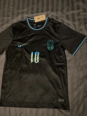 Svart Brasilien fotbollströja Nike neymar jr 10 - Ej använd med tagg. Snygg svart fotbollströja från Nike med Brasilien-emblem och nummer 10 neymar jr i turkos. Tröjan har turkosa detaljer vid krage och ärmslut, samt Nike-logga på bröstet. Perfekt för dig som älskar fotboll och vill sticka ut på planen.