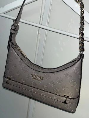  axelväska från Guess - Snygg handväska från Guess med präglat logomönster och guldfärgade detaljer. Väskan har en axelrem i kombinerat läder och kedja, samt dragkedja upptill. Perfekt storlek för det viktigaste och ger en trendig touch till din outfit.
