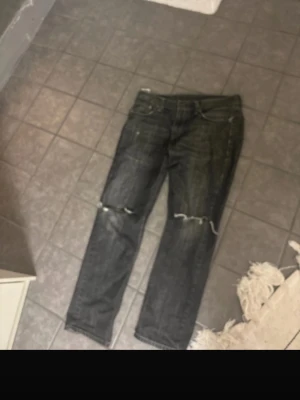Svarta slitna jeans med hål - Säljer ett par svarta jeans med raka ben och slitna detaljer, inklusive hål på båda knäna. Jeansen har klassisk femficksmodell och är tillverkade i denim. Perfekta för en avslappnad och trendig look.