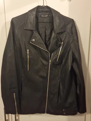 Svart skinnjacka Soul River - Snygg svart skinnjacka från Soul River med klassisk bikerstil. Jackan har snedställd dragkedja, tre framfickor med dragkedjor och detaljer vid ärmsluten. Perfekt för dig som vill ha en tidlös och cool look.