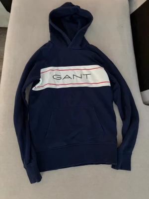 Marinblå hoodie från GANT - Snygg marinblå hoodie från GANT med stor vit panel och röda linjer över bröstet samt GANT-logga tryckt framtill. Klassisk känguruficka och ribbade muddar. 