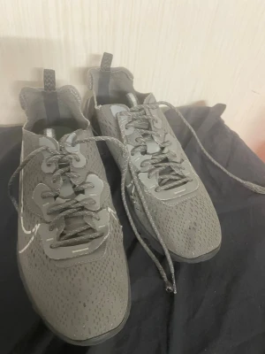 Nike grå sneakers med mesh - Säljer ett par grå Nike sneakers med coolt meshmaterial och reflexdetaljer. Skorna har klassisk låg profil, snörning och en tydlig vit swoosh på sidan. Perfekta för dig som gillar sportig och modern stil.