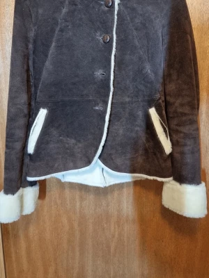 Brun pälsjacka med huva - Mysig brun pälsjacka med vit fuskpäls på insidan, vid ärmslut och fickor. Jackan har knäppning framtill, två snedställda fickor och en huva. Perfekt för kalla vinterdagar och ger en cool vintagekänsla.