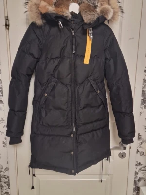 Svart Parajumpers parkas med päls - Snygg svart vinterjacka från Parajumpers med stor huva och äkta pälsdetalj. Jackan har dragkedja, knappar, stora fickor och klassisk gul rem vid halsen. Perfekt för kalla dagar och riktigt varm. Märkespatch på ärmen. 💞pris kan diskuteras men 5000 är rimligt då nypris är 10.000