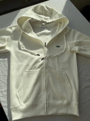 Vit zip hoodie från Lacoste - Snygg vit hoodie från Lacoste med dragkedja framtill och klassisk krokodil-logga på bröstet. Hoodien har stor huva, känguruficka och ribbade muddar. Tillverkad i mjuk bomull som känns skön mot huden. Perfekt för en clean och stilren look.