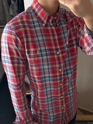 Rutig skjorta från Polo Ralph Lauren - Riktigt snygg rutig skjorta från Polo Ralph Lauren i rött, blått och vitt. Klassisk button-down krage, långärmad och med broderad gul logga på bröstet. Skjortan är i mjuk bomull och har knappar framtill.