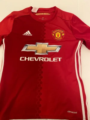 Manchester United matchtröja Adidas - Röd Manchester United matchtröja från Adidas med vitt tryck och guldigt Chevrolet-märke på bröstet. Kortärmad modell i lätt och ventilerande polyester med klubbmärke och Adidas-logga. Perfekt för fotbollsträning eller match.
