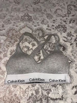 Grå sport-bh från Calvin Klein - Säljer en grå sport-bh från Calvin Klein med korsade axelband i ryggen och bred resår med logga. Bh:n är mjuk och utan bygel, tillverkad i stretchigt material för extra komfort. Perfekt för en aktiv vardag eller som skön lounge-bh. Har en i svart också som jag inte lagt upp ännu! 