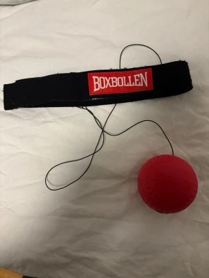 Boxbollen träningsboll med pannband - Säljer en Boxbollen träningsboll med svart elastiskt pannband och röd boll i mjukt material. Perfekt för reflex- och boxningsträning. Bollen sitter fast i ett snöre som är fäst i pannbandet. Enkel att använda för att träna koordination och snabbhet.