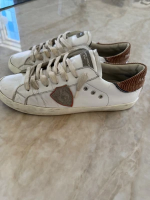 Philippe Model vita sneakers med brunt - Snygga vita sneakers från Philippe Model med brunt ribbat parti på hälen och grå detaljer. Skorna har klassisk låg profil, vita snören och ett emblem på sidan. Materialet är läder med textil på hälen. De är snygga och box följer tyvärr inte med där av det låga priset🔥👟