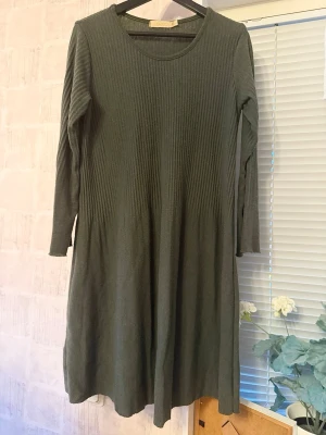 Ullklänning - Klänningen är i en djup mörkgrön nyans och har en smickrande ribbstickad textur upptill som övergår i ett mjukt fall. Stretchigt material.  Storlek: L/XL (flexibel passform tack vare stretchen) Längd: ca 93 cm Material: 60% viskos, 35% ull, 5% elastan Skick: Klänningen är något nopprig (se bilder), men i övrigt i fint skick. Nopporna går enkelt att åtgärda med en noppborttagare.