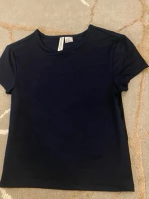 Mörkblå basic t-shirt från H&M - En enkel och stilren mörkblå t-shirt från H&M med rund halsringning och korta ärmar. Perfekt att matcha med jeans eller kjol för en clean look. Tillverkad i mjuk bomull för skön känsla hela dagen.