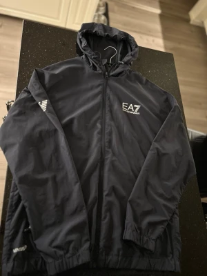 Svart EA7 Emporio Armani vindjacka - Snygg svart vindjacka från EA7 Emporio Armani med huva och dragkedja framtill. Jackan har vita logotyper på bröstet och ärmen, samt elastiska muddar vid ärmslut. Perfekt för en sportig och stilren look.KOM MED BUD, OBS FLÄCKAR (går att ta bort)