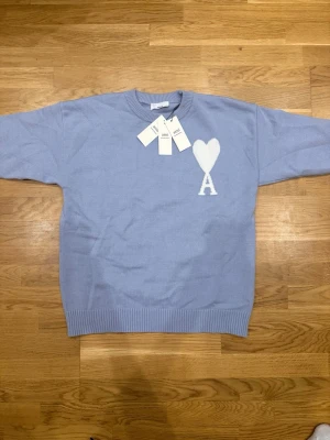 Blå stickad tröja från Ami - Snygg ljusblå stickad tröja från Ami med ett stort vitt hjärta och bokstaven A broderat på bröstet. Tröjan har rund halsringning och ribbstickade muddar vid ärmslut och nederkant. Perfekt för dig som gillar stilrena detaljer.