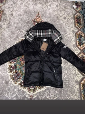 Burberry jacka - Snygg svart pufferjacka från Burberry med klassiskt rutigt foder i huvan och tydlig logotyp på ärmen. Storlek M men passar S, pris kan diskuteras!