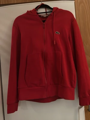 Röd hoodie från Lacoste med dragkedja storlek Small - Storlek Small i fint skick! Snygg röd hoodie från Lacoste med dragkedja framtill och klassisk krokodillogga på bröstet. Hoodien har två stora fickor fram och justerbar huva med snören. Tillverkad i mjukt bomullsmaterial som känns skönt mot huden. Perfekt för en chill och sportig look. Väldigt lite använd då den va för gränsen för liten