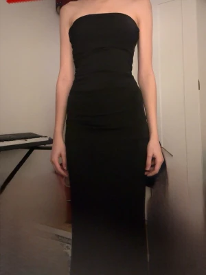 Svart tub långklänning - Säljer en elegant svart axelbandslös långklänning med figurnära passform. Klänningen har en enkel och stilren design utan mönster eller detaljer, perfekt för dig som gillar minimalistiskt mode. Materialet känns mjukt och följsamt mot kroppen.     Använt fåtal gånger 