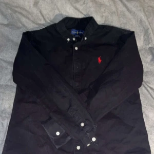 Svart skjorta från Ralph Lauren - Klassisk svart skjorta från Ralph Lauren med röd broderad logga på bröstet. Skjortan har knappar framtill och på kragen samt långa ärmar. Tillverkad i bomull för en skön känsla och stilren look. Den är Storlek L i barnstorlek, motsvarar ungefär S i Vuxenstorlek