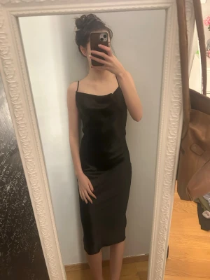 Svart långklänning med snörning - Svart långklänning med smala axelband och öppen rygg med snörningsdetalj. Klänningen har en slank passform och är tillverkad i ett glansigt material som ger en elegant look. Perfekt för dig som vill ha en stilren och trendig klänning.