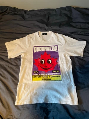 Vit Dsquared2 t-shirt med tryck - Vit Dsquared2 T-shirt i storlek 14 år men sitter som S/XS. Nytt skick endast använd ett fåtal gånger.