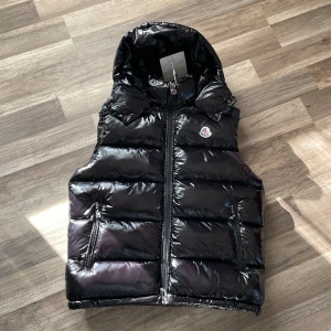 Moncler vest  - Snygg svart dunväst från Moncler. Västen har en broderad logotyp på bröstet och praktiska fickor med dragkedja. Perfekt för kyligare dagar.