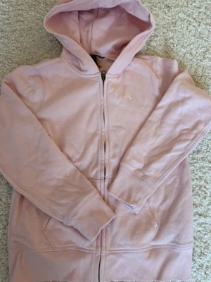 Ljusrosa hoodie från Polo Ralph Lauren - Snygg ljusrosa hoodie med dragkedja från Polo Ralph Lauren. Klassisk design med huva, fickor framtill och den ikoniska vita logon broderad på bröstet. Perfekt för en avslappnad och trendig look. Det är XL i  BARNSTORLEK. Nypris 1300