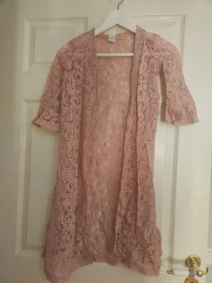 Rosa spetskofta med trumpetärm - Säljer en ljusrosa, lång spetskofta med trekvarts trumpetärmar. Koftan är öppen framtill och har ett vackert blommigt spetsmönster som ger en romantisk vibe. Perfekt att slänga över en topp för en extra touch.