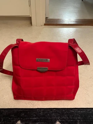 Röd axelväska från Carnaby - Snygg röd axelväska från Carnaby med quiltad design och justerbar axelrem. Väskan har ett lock med metallogo framtill och dragkedjefack på baksidan. Materialet är en mix av textil och konstläder vilket ger en modern look.