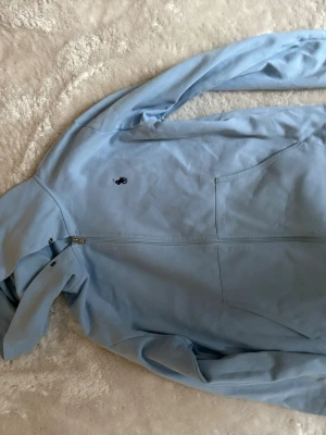 Ljusblått loungewear-set från Polo Ralph Lauren - Mysigt loungewear-set i ljusblått från Polo Ralph Lauren. Setet består av en hoodie med dragkedja och fickor samt matchande mjukisbyxor med resår i midjan och mudd vid benslut. Båda plaggen har den klassiska Polo-loggan broderad på bröstet och benet.