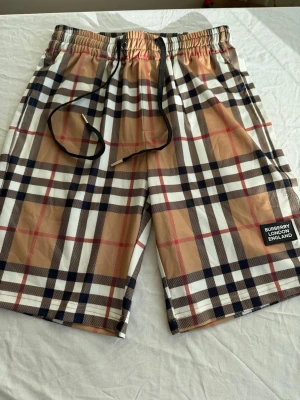 Rutig Burberry shorts med snörning - Säljer ett par klassiska Burberry shorts i beige, svart, vit och röd med det ikoniska rutiga mönstret. Shortsen har elastisk midja med svart snörning och en patch med Burberry London England på benet. Perfekta för sommaren och riktigt snygga till streetwear.
