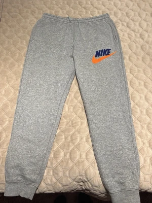 Gråa mjukisbyxor från Nike - Snygga gråa mjukisbyxor från Nike med blå logga och orange swoosh på vänster lår. Byxorna har resår och dragsko i midjan samt muddar vid bensluten. Perfekta för chill eller träning, tillverkade i mjukt material.