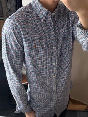 Rutig skjorta från Polo Ralph Lauren - Klassisk rutig skjorta från Polo Ralph Lauren i bomull. Skjortan har blå, röda och gröna rutor, button-down krage och långa ärmar. Ikoniska broderade loggan på bröstet. Perfekt för en stilren och avslappnad look. Lite missfärgad krage men syns knappt 