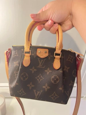 Louis Vuitton Monogram handväska - Säljer en klassisk Louis Vuitton handväska i brunt monogrammönster med beige läderhandtag och guldfärgade metalldetaljer. Väskan har dragkedja upptill och avtagbar axelrem. Perfekt storlek för det viktigaste och ikonisk LV-design.