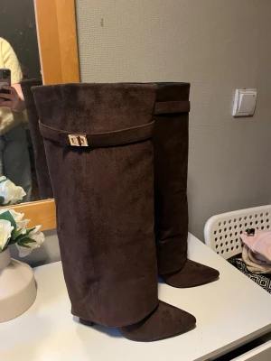 Bruna höga mockastövlar med spänne - Snygga bruna stövlar i mockaimitation med spetsig tå och dekorativt guldfärgat spänne upptill. Stövlarna är höga och har en bredare skaftform som ger en cool och trendig look. Perfekta till höstens outfits och passar till både jeans och kjol.